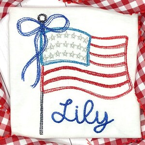 K&ouml;nnte beinhalten: Wei&szlig;er Stoff mit einem gestickten amerikanischen Flaggen-Design. Die Flagge hat rote und wei&szlig;e Streifen, ein blaues Feld mit silbernen Sternen und eine blaue Schleife. Der Name "Lily" ist darunter in blauer Schrift gestickt.