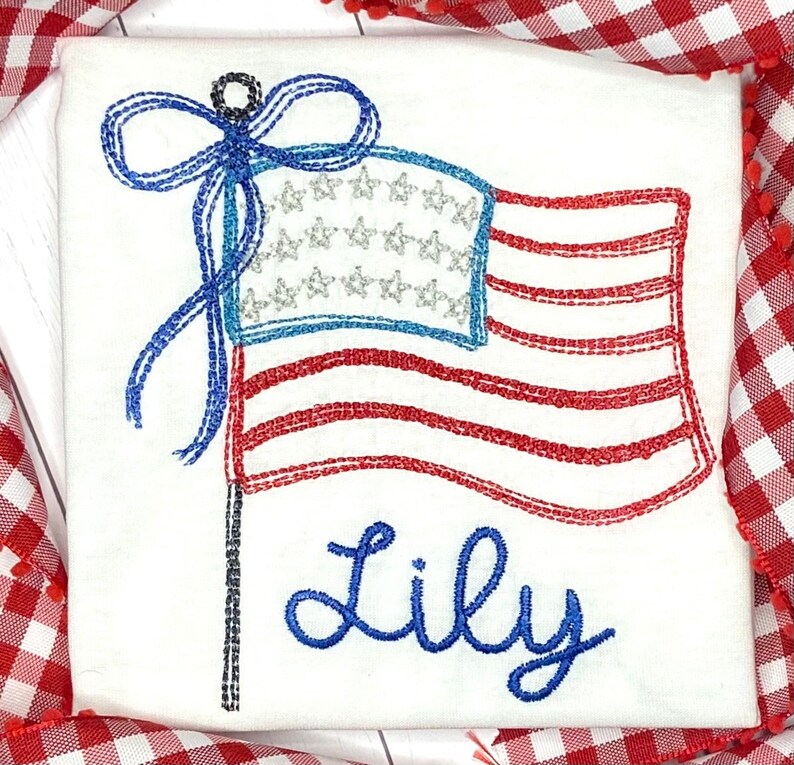 K&ouml;nnte beinhalten: Wei&szlig;er Stoff mit einem gestickten amerikanischen Flaggen-Design. Die Flagge hat rote und wei&szlig;e Streifen, ein blaues Feld mit silbernen Sternen und eine blaue Schleife. Der Name "Lily" ist darunter in blauer Schrift gestickt.
