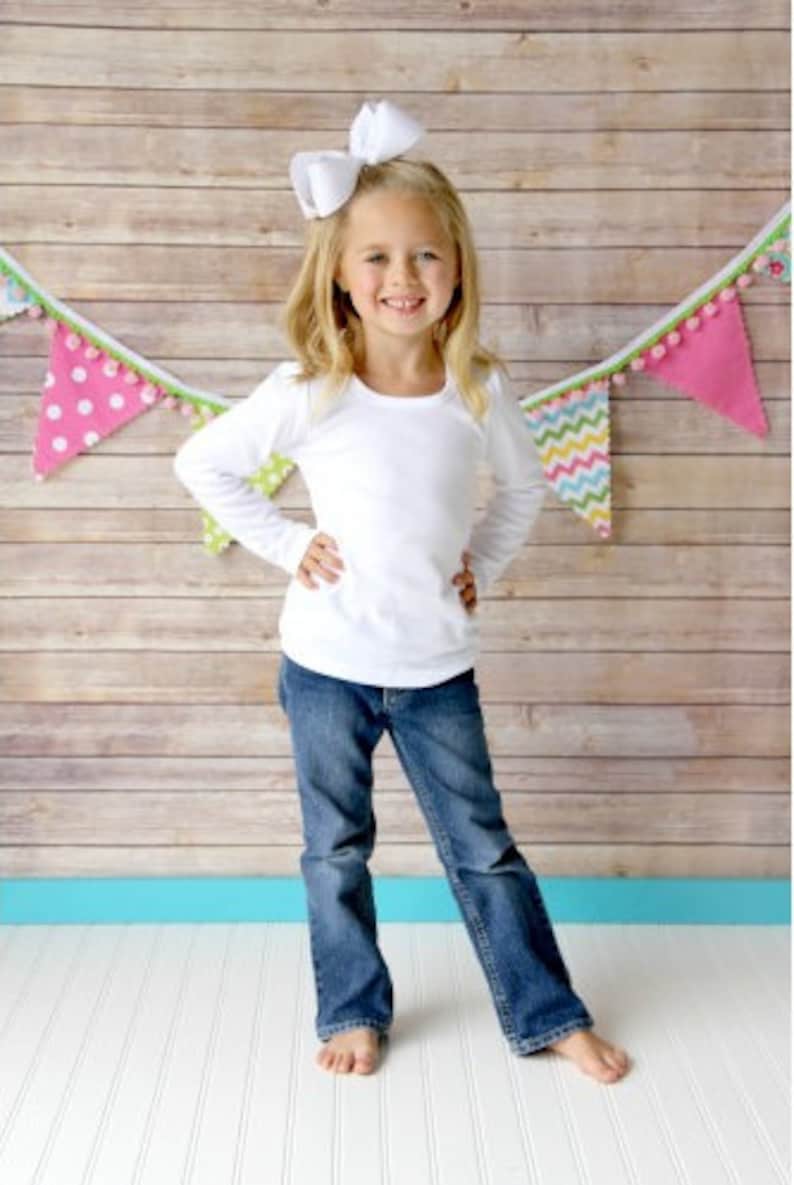 Puede incluir: Una ni&ntilde;a peque&ntilde;a con una camisa blanca de manga larga y pantalones vaqueros azules, con un lazo blanco en el pelo, de pie frente a una pared de madera con una pancarta colorida colgada encima de ella.