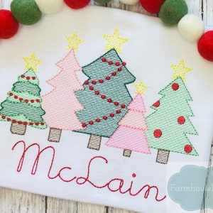 May include: A white t-shirt with an embroidered design of five Christmas trees with red ornaments and lights. The trees are in different colors, including green, pink, and blue. The name "McClain" is embroidered below the trees.