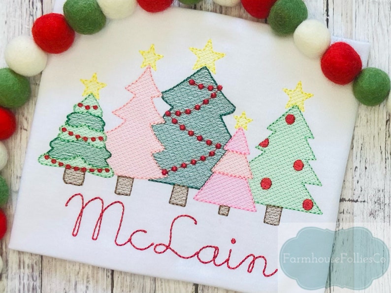 May include: A white t-shirt with an embroidered design of five Christmas trees with red ornaments and lights. The trees are in different colors, including green, pink, and blue. The name "McClain" is embroidered below the trees.