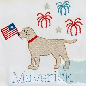 Op de afbeelding: Een wit geborduurd T-shirt met een gele labrador retriever die een Amerikaanse vlag vasthoudt. Het shirt heeft ook rood, wit en blauwe vuurwerk en sterren. De naam "Maverick" is onder de hond geborduurd.