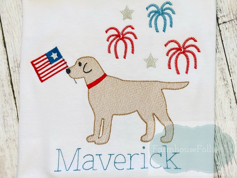 Op de afbeelding: Een wit geborduurd T-shirt met een gele labrador retriever die een Amerikaanse vlag vasthoudt. Het shirt heeft ook rood, wit en blauwe vuurwerk en sterren. De naam "Maverick" is onder de hond geborduurd.
