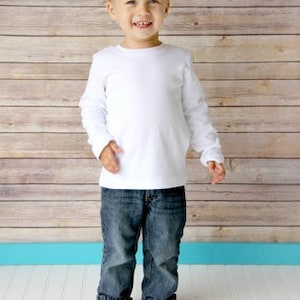 May include: A young boy with spiky blond hair is wearing a white long-sleeved shirt and blue jeans. He is standing in front of a wood-paneled wall.