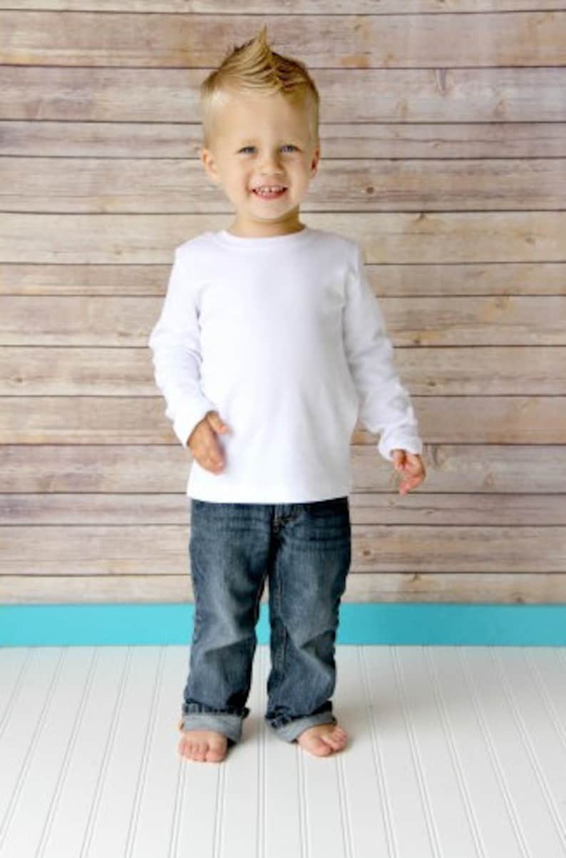 May include: A young boy with spiky blond hair is wearing a white long-sleeved shirt and blue jeans. He is standing in front of a wood-paneled wall.