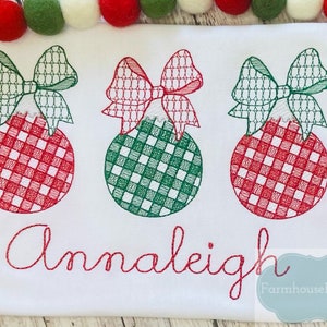 Pode incluir: Tr&ecirc;s enfeites de Natal vermelhos e verdes com la&ccedil;os, bordados em um tecido branco com o nome "Annaleigh" e o texto "Farmhouse Follies Co."