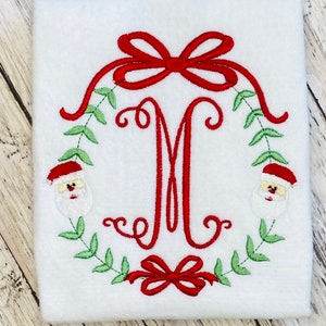 Puede incluir: Guirnalda navide&ntilde;a bordada con un lazo rojo y caras de Pap&aacute; Noel a cada lado de la letra M.