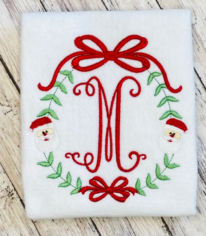 Puede incluir: Guirnalda navide&ntilde;a bordada con un lazo rojo y caras de Pap&aacute; Noel a cada lado de la letra M.