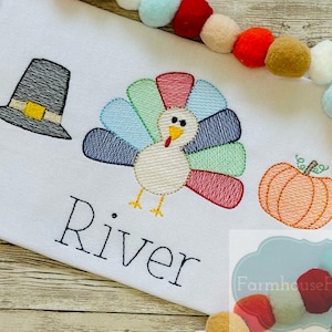 Pode incluir: Uma camiseta branca com um desenho bordado de um chapéu de peregrino, um peru e uma abóbora. O nome "River" está bordado abaixo do desenho.