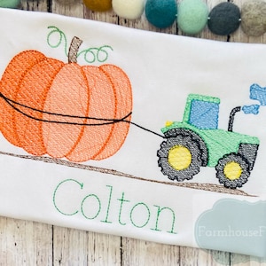 May include: Embroidered design of a green tractor pulling an orange pumpkin with the name Colton in green lettering below. The design is on a white t-shirt.
