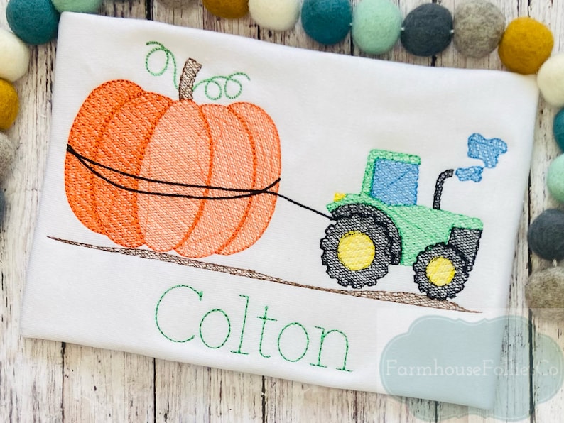 May include: Embroidered design of a green tractor pulling an orange pumpkin with the name Colton in green lettering below. The design is on a white t-shirt.
