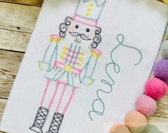 May include: Embroidered white shirt with a colorful nutcracker design and the name "Ava" embroidered in cursive.