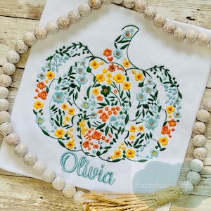 May include: White t-shirt featuring an embroidered floral pumpkin design and the name "Olivia" in teal lettering.