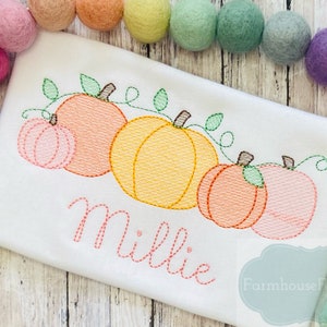 May include: A white onesie with an embroidered design of four pumpkins with green vines and leaves. The name "Millie" is embroidered below the pumpkins. The onesie is surrounded by colorful pom-poms.
