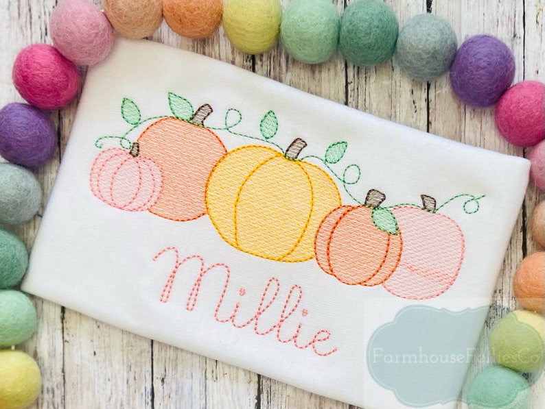 May include: A white onesie with an embroidered design of four pumpkins with green vines and leaves. The name "Millie" is embroidered below the pumpkins. The onesie is surrounded by colorful pom-poms.