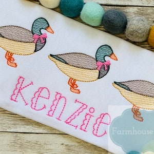 Puede incluir: Una tela blanca con tres patos bordados que llevan lazos rosas. Los patos est&aacute;n en fila y el nombre "Kenzie" est&aacute; bordado debajo de ellos en hilo rosa. El texto "Farmhouse Follies" tambi&eacute;n est&aacute; bordado en hilo azul.
