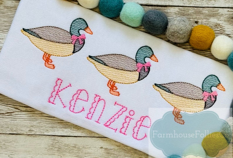 Puede incluir: Una tela blanca con tres patos bordados que llevan lazos rosas. Los patos est&aacute;n en fila y el nombre "Kenzie" est&aacute; bordado debajo de ellos en hilo rosa. El texto "Farmhouse Follies" tambi&eacute;n est&aacute; bordado en hilo azul.