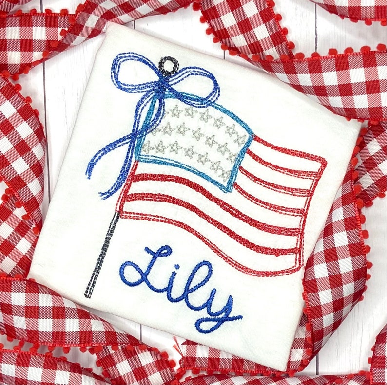 K&ouml;nnte beinhalten: Wei&szlig;es T-Shirt mit einem gestickten Design der amerikanischen Flagge. Die Flagge hat rote und wei&szlig;e Streifen, ein blaues Feld mit silbernen Sternen und eine blaue Schleife. Der Name "Lily" ist darunter in blauer Schrift gestickt.