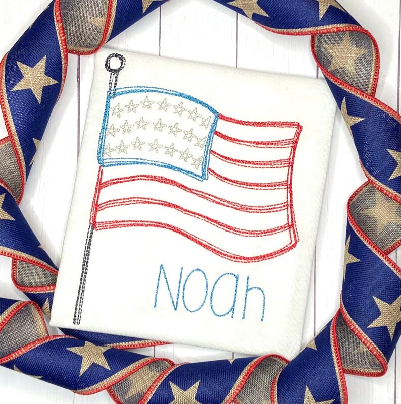May include: White embroidered t-shirt with a red, white, and blue American flag and the name "Noah" in blue thread.