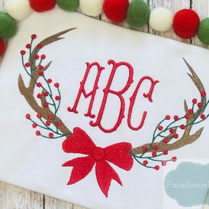 May include: White embroidered shirt with a red bow, brown antlers, and green and red berries. The monogram "ABC" is embroidered in red.
