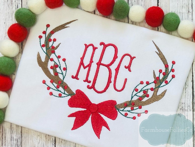 May include: White embroidered shirt with a red bow, brown antlers, and green and red berries. The monogram "ABC" is embroidered in red.