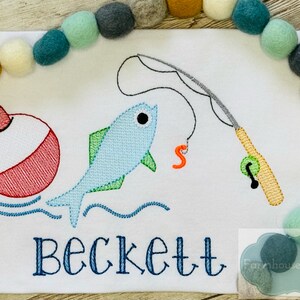 Puede incluir: Dise&ntilde;o bordado de un pez azul, un flotador rojo y blanco y una ca&ntilde;a de pescar con un flotador verde. El nombre Beckett est&aacute; bordado debajo del dise&ntilde;o.