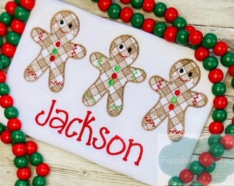 May include: White t-shirt featuring three gingerbread man designs with brown, red, and green accents. The name "Jackson" is embroidered in red script below the gingerbread men. Red and green bead garland frames the shirt.
