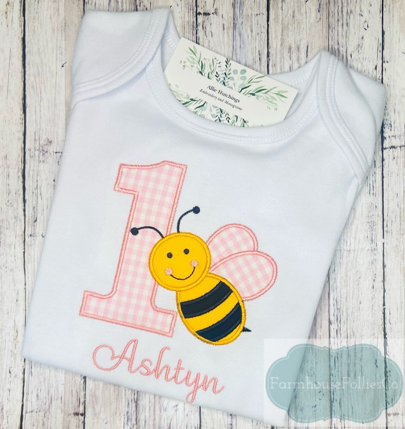 May include: White onesie with a pink and white gingham number one and a yellow and black embroidered bee. The onesie is personalized with the name "Ashtyn" in pink.