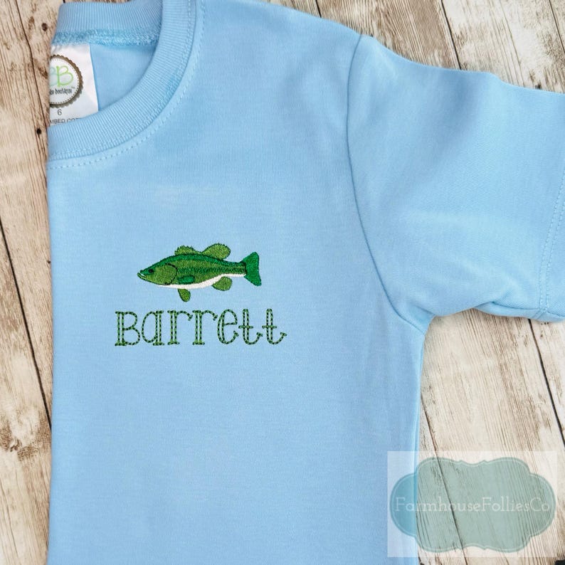 May include: A light blue short-sleeved t-shirt with a green embroidered fish and the name "Barrett" in green thread. The shirt has a round neckline and a tag on the upper left side.