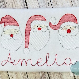 May include: White fabric with three embroidered Santa faces and the name Amelia in red thread.