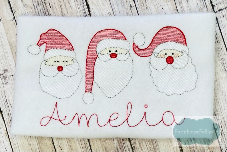 May include: White fabric with three embroidered Santa faces and the name Amelia in red thread.