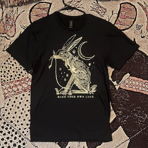 May include: Black t-shirt with a gold graphic of a rabbit holding a string with a crescent moon and stars in the background. The text "Make your own luck" is printed below the rabbit.