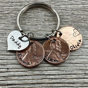 May include: A silver heart charm with the word "Daddy" engraved on it, attached to a silver key ring with four copper pennies. Each penny has a heart engraved on it with a year and a name: "Emma" 2012, "Liam" 2015, and "Olivia" 2020.