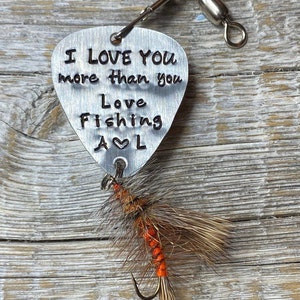 May include: A silver guitar pick with the text "I love you more than you Love Fishing A♥L" attached to a brown and orange fishing lure.