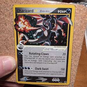May include: A Charizard Delta Species Pokémon card with a yellow border. The card features a black and orange illustration of Charizard breathing fire. Text includes "Charizard," "90HP," and attack descriptions. The card is from 2006.