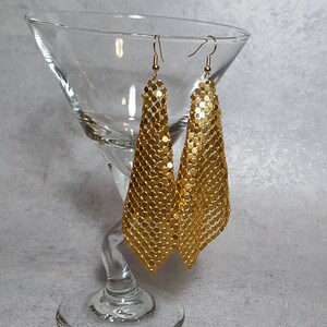 May include: A pair of gold-colored, dangling earrings with a chainmail design. The earrings are displayed in a clear martini glass against a gray background. The earrings have a triangular shape and are made of small, connected metal pieces.