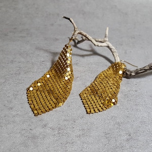 May include: A pair of gold-toned dangle earrings made of a chainmail-like material. The earrings are hanging from a branch.