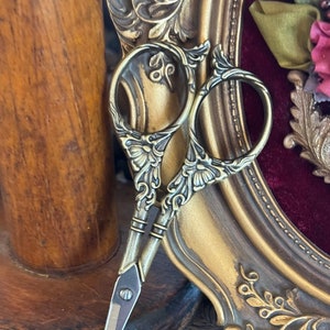 Könnte beinhalten: Verzierte goldfarbene Schere mit floralem Design, die auf einer Holzoberfläche und einem goldenen Bilderrahmen liegt.