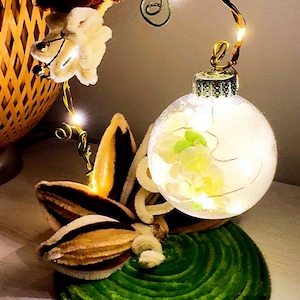 May include: A decorative lamp featuring a clear glass sphere filled with white flowers and fairy lights, suspended from a curved wire arm. The base is a circular green coiled material, accented with brown and white fabric petals and floral elements.