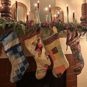 May include: A fireplace mantel decorated for the holidays. Four quilted Christmas stockings hang below a garland of greenery and red accents. Lit candles and decorative candle holders are on the mantel.