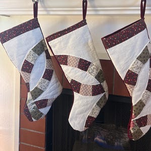 May include: Three quilted Christmas stockings featuring a white fabric base with a decorative patchwork pattern of interlocking rings in shades of burgundy, olive green, and cream floral prints. Each stocking has a dark burgundy fabric cuff and loop.
