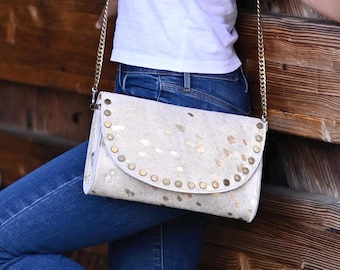 Könnte beinhalten: Eine Frau in einem weißen Tanktop und blauen Jeans steht an einer Holzwand. Sie trägt eine weiße Leder-Umhängetasche mit goldenen Nieten.