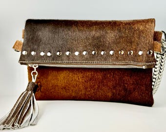 Może przedstawiać: Brązowa i szara torebka typu clutch ze skóry krowiej z srebrnym łańcuszkiem i srebrnym frędzlem. Torebka ma zamek błyskawiczny i jest ozdobiona srebrnymi ćwiekami.