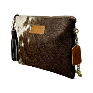 K&ouml;nnte beinhalten: Braune und wei&szlig;e Kuhfell-Clutch mit goldener Kettenkette und braunem Leder-Quaste. Die Tasche hat ein braunes Lederetikett mit dem Text "SHANE TRIFID" darauf.