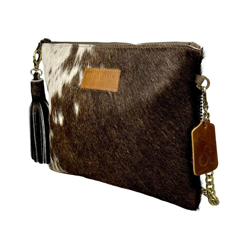 K&ouml;nnte beinhalten: Braune und wei&szlig;e Kuhfell-Clutch mit goldener Kettenkette und braunem Leder-Quaste. Die Tasche hat ein braunes Lederetikett mit dem Text "SHANE TRIFID" darauf.