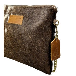K&ouml;nnte beinhalten: Eine braune und wei&szlig;e Clutch aus Kuhfell mit einem braunen Lederriemen und einer goldenen Kette. Die Tasche hat ein braunes Leder-Etikett mit dem Text "Shane Thread" und einen goldenen Anh&auml;nger mit den Initialen "ST".