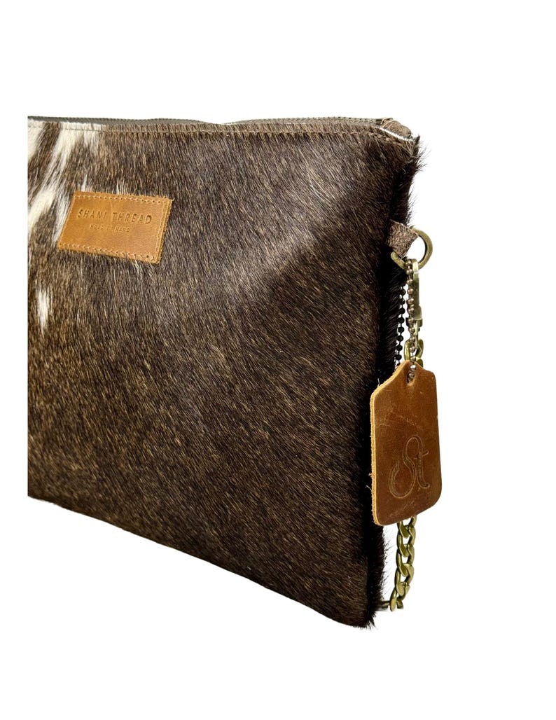 K&ouml;nnte beinhalten: Eine braune und wei&szlig;e Clutch aus Kuhfell mit einem braunen Lederriemen und einer goldenen Kette. Die Tasche hat ein braunes Leder-Etikett mit dem Text "Shane Thread" und einen goldenen Anh&auml;nger mit den Initialen "ST".