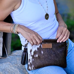 K&ouml;nnte beinhalten: Eine Frau tr&auml;gt ein wei&szlig;es Tanktop und eine blaue Jeans und h&auml;lt eine braune und wei&szlig;e Kuhfell-Clutch mit einem schwarzen Quastenanh&auml;nger. Die Tasche hat ein braunes Lederetikett mit der Aufschrift "BRAN + TABARD".