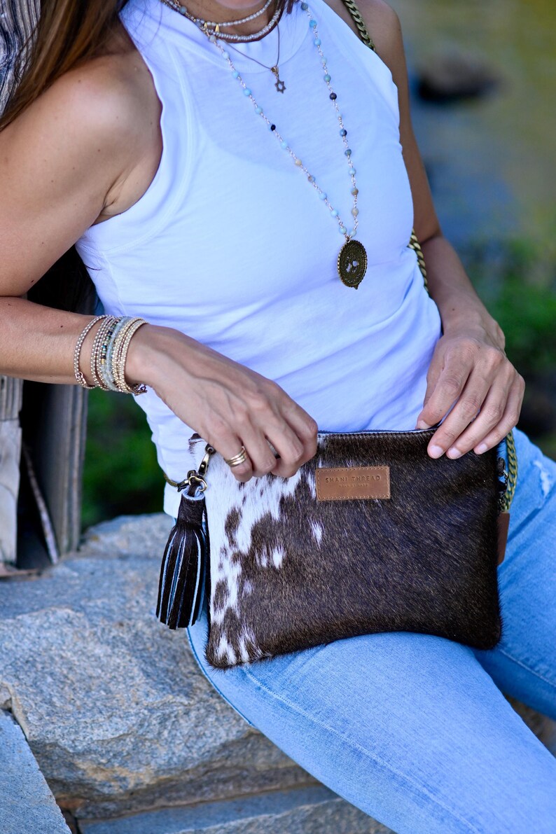 K&ouml;nnte beinhalten: Eine Frau tr&auml;gt ein wei&szlig;es Tanktop und eine blaue Jeans und h&auml;lt eine braune und wei&szlig;e Kuhfell-Clutch mit einem schwarzen Quastenanh&auml;nger. Die Tasche hat ein braunes Lederetikett mit der Aufschrift "BRAN + TABARD".