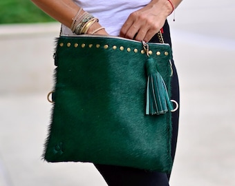 Könnte beinhalten: Eine dunkelgrüne, flauschige Ledertasche mit goldenen Akzenten und einem Quastenanhänger. Die Tasche hat einen Reißverschluss und einen langen Riemen.
